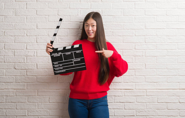 Young Chinese woman over white brick wall holding clapboard very happy pointing with hand and finger - Foto, imagen