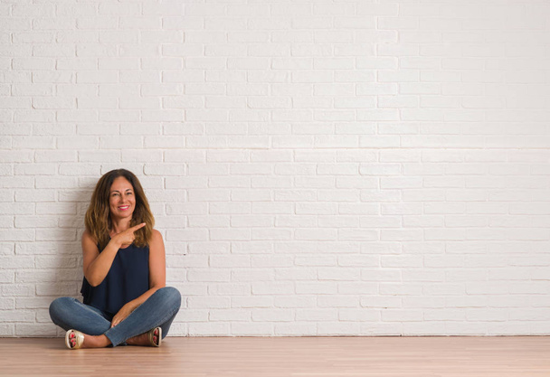 Middle age hispanic woman sitting on the floor over white brick wall cheerful with a smile of face pointing with hand and finger up to the side with happy and natural expression on face looking at the camera. - Photo, Image
