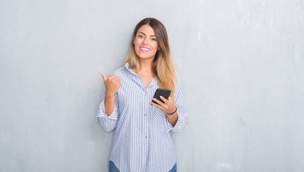 Young adult woman over grey grunge wall looking at smartphone texting a message pointing and showing with thumb up to the side with happy face smiling - Φωτογραφία, εικόνα