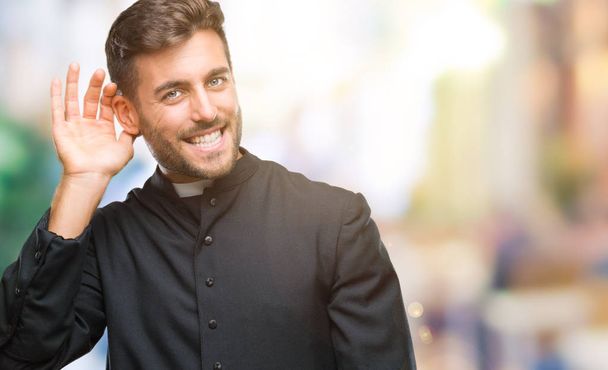junger katholischer christlicher Priester mit isoliertem Hintergrund, der lächelnd mit der Hand über dem Ohr Gerüchten oder Klatsch lauscht. Konzept für Gehörlosigkeit. - Foto, Bild