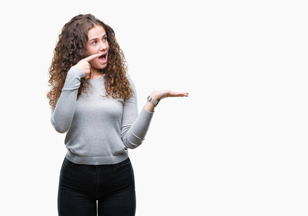 Beautiful brunette curly hair young girl wearing a sweater over isolated background amazed and smiling to the camera while presenting with hand and pointing with finger. - Photo, Image
