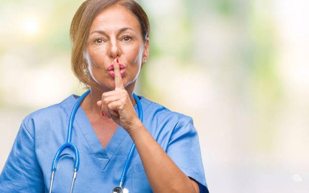Μέση ηλικία ανώτερος νοσοκόμα γιατρός γυναίκα πέρα από το απομονωμένο υπόβαθρο ζητώντας να είναι ήσυχη με το δάχτυλο στα χείλη. Σιωπή και μυστικό έννοια. - Φωτογραφία, εικόνα