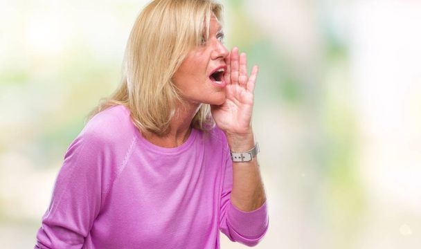Blonde vrouw van middelbare leeftijd op geïsoleerde achtergrond schreeuwen en schreeuwen luid naar de andere kant met hand op de mond. Communicatieconcept. - Foto, afbeelding