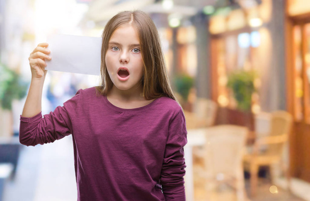 Menina bonita jovem segurando cartão de papel em branco sobre fundo isolado assustado em choque com um rosto surpresa, com medo e animado com expressão de medo
 - Foto, Imagem