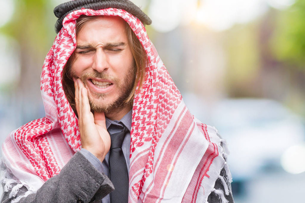 Nuori komea arabialainen mies, jolla on pitkät hiukset yllään keffiyeh yli eristetty tausta koskettaa suuta käsin kivulias ilme, koska hammassärky tai hampaiden sairaus hampaissa. Hammaslääkärin käsite
. - Valokuva, kuva