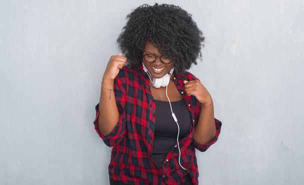Молодая африканская американка через серую гранж-стену в наушниках очень счастлива и взволнована, делая жест победителя с поднятыми руками, улыбаясь и крича об успехе. Концепция празднования
. - Фото, изображение