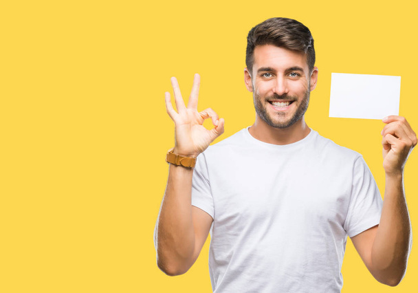 Young handsome man holding blank card over isolated background doing ok sign with fingers, excellent symbol - Zdjęcie, obraz