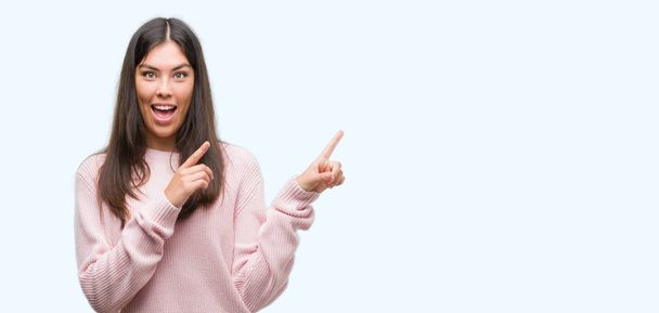 Jovem mulher hispânica bonita vestindo uma camisola sorrindo e olhando para a câmera apontando com duas mãos e dedos para o lado
. - Foto, Imagem