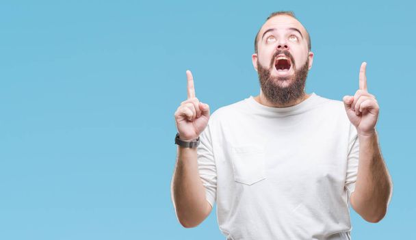 Young caucasian hipster man wearing casual t-shirt over isolated background amazed and surprised looking up and pointing with fingers and raised arms. - Photo, Image