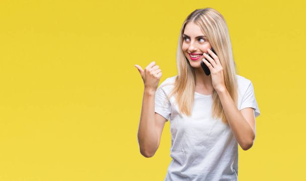 Young beautiful blonde woman talking using smartphone over isolated background pointing and showing with thumb up to the side with happy face smiling - Fotografie, Obrázek
