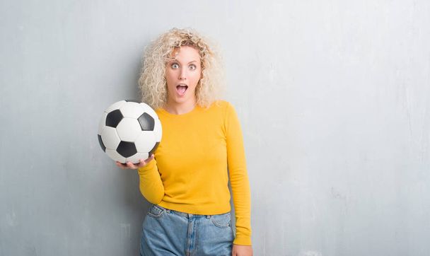 Young blonde woman over grunge grey background holding soccer football ball scared in shock with a surprise face, afraid and excited with fear expression - Valokuva, kuva