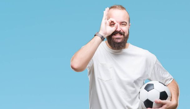 Giovane uomo hipster caucasico che tiene palla da calcio su sfondo isolato con volto felice sorridente facendo ok segno con mano sugli occhi guardando attraverso le dita
 - Foto, immagini
