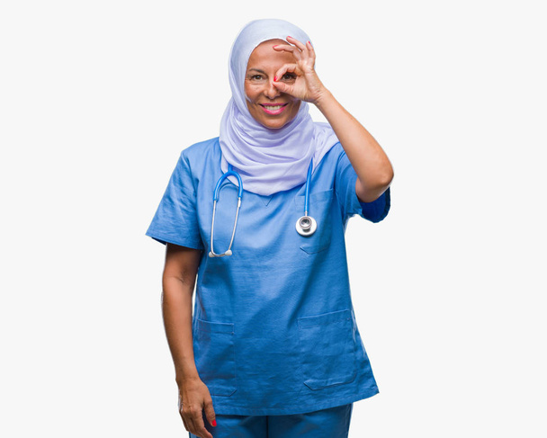 Enfermera árabe mayor de mediana edad que usa hijab sobre un fondo aislado haciendo un gesto bien con la mano sonriente, ojos mirando a través de los dedos con cara feliz
. - Foto, imagen