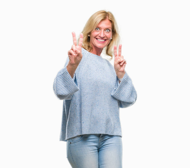 Μέσης ηλικίας ξανθιά γυναίκα, φορώντας χειμώνα πουλόβερ πάνω από απομονωμένες φόντο χαμογελώντας ψάχνει στην κάμερα δείχνει τα δάχτυλα που κάνει το σήμα της νίκης. Αριθμός δύο. - Φωτογραφία, εικόνα