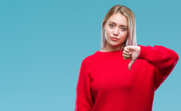 若いブロンドの女性は不幸なと怒って拒否反応を示さず、ジェスチャーを親指で否定的な分離の背景に冬のセーターを着ています。不適切な表現. - 写真・画像