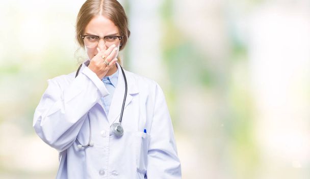 Όμορφο Νεαρό ξανθιά γιατρός γυναίκα φορώντας ιατρική στολή πάνω από τα απομονωμένα φόντο μυρίζουν κάτι stinky και αηδιαστικό, αφόρητη μυρωδιά, εκμετάλλευση αναπνοή με δάκτυλα στη μύτη. Άσχημες μυρωδιές έννοια. - Φωτογραφία, εικόνα