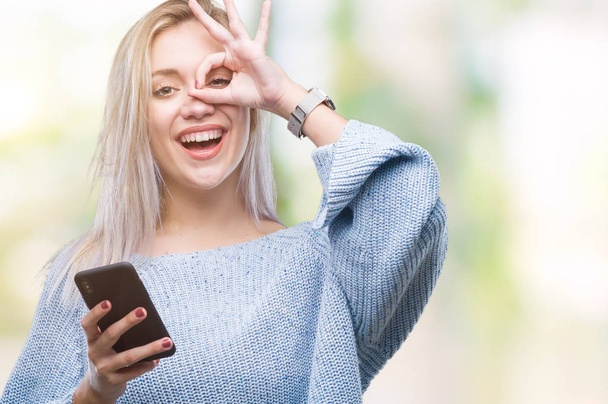 指の間から見て目に手で ok サインをして笑っている幸せそうな顔で孤立の背景にスマート フォンを使用してメッセージを送信する若いブロンドの女性のテキスト メッセージ - 写真・画像