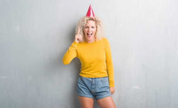 Sarışın genç kadın Doğum günü şapkası işaret parmağı ile başarılı fikir giyen grunge gri duvarın üzerinden. Çıktıktan ve mutlu. Bir numara. - Fotoğraf, Görsel