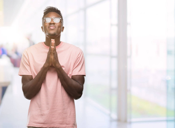 Jeune homme afro-américain portant un t-shirt rose priant les mains jointes demandant pardon en souriant confiant
. - Photo, image
