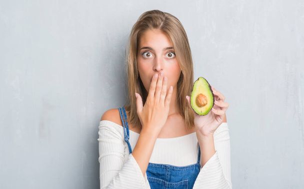 Beautiful young woman over grunge grey wall eating avocado cover mouth with hand shocked with shame for mistake, expression of fear, scared in silence, secret concept - Photo, Image