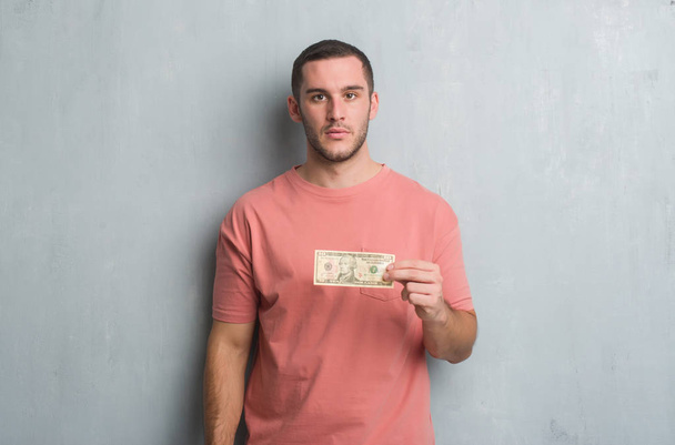 Jeune homme caucasien sur le mur gris grunge montrant dix dollars avec une expression confiante sur la pensée intelligente visage grave
 - Photo, image