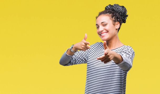 Молодий плетені волосся афро-американських дівчата носити светр над ізольованих фону, вказуючи пальцем камери з щасливим і смішна обличчя. Хорошу енергію і вібрації. - Фото, зображення