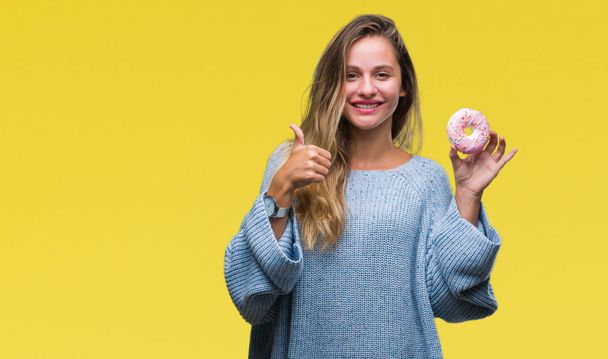 Jovem bela mulher loira comendo donut doce sobre fundo isolado feliz com grande sorriso fazendo ok sinal, polegar para cima com os dedos, excelente sinal
 - Foto, Imagem