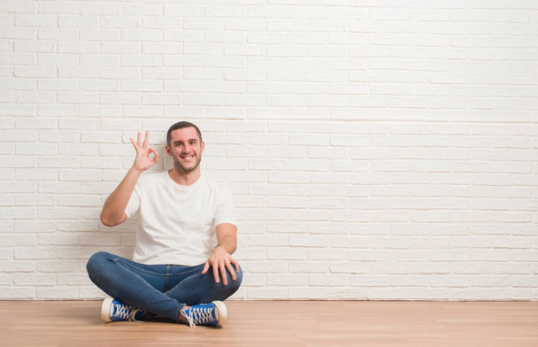 Homem caucasiano jovem sentado no chão sobre a parede de tijolo branco sorrindo positivo fazendo sinal ok com a mão e os dedos. Expressão bem sucedida
. - Foto, Imagem