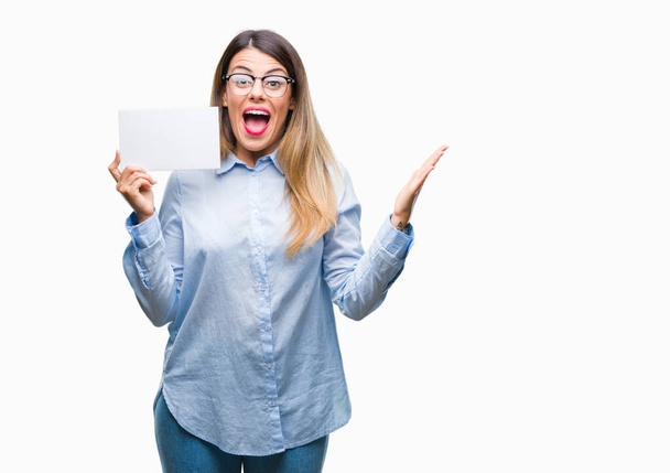 Young beautiful business woman holding blank card over isolated background very happy and excited, winner expression celebrating victory screaming with big smile and raised hands - Φωτογραφία, εικόνα