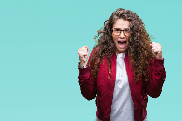 Beautiful brunette curly hair young girl wearing jacket and glasses over isolated background excited for success with arms raised celebrating victory smiling. Winner concept. - Foto, Imagen