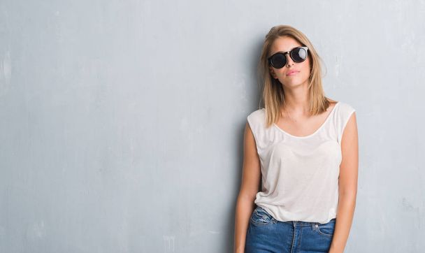 Hermosa mujer joven de pie sobre la pared gris grunge usando gafas de sol de moda con una expresión segura en la cara inteligente pensando en serio
 - Foto, imagen