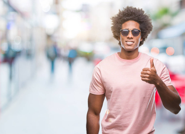 Hombre afroamericano con gafas de sol sobre un fondo aislado haciendo un gesto feliz con la mano. Aprobar la expresión mirando a la cámara con éxito
. - Foto, imagen