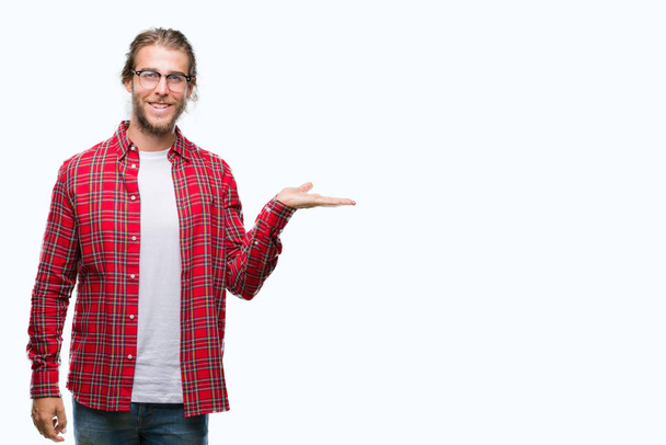 Mladý pohledný muž s dlouhými vlasy, nosí brýle izolované pozadí s úsměvem veselá prezentaci a polohovací s dlaň ruky při pohledu na fotoaparát. - Fotografie, Obrázek