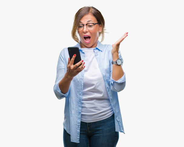 Seniorin mittleren Alters sendet SMS mit Smartphone über isolierten Hintergrund sehr glücklich und aufgeregt, Siegerausdruck feiert Sieg schreiend mit breitem Lächeln und erhobenen Händen - Foto, Bild