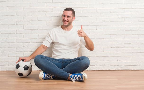 Молодий білий чоловік сидить над білою цегляною стіною, тримає футбольний м'яч щасливим з великою посмішкою роблячи знак ок, великий палець пальцями, відмінний знак
 - Фото, зображення