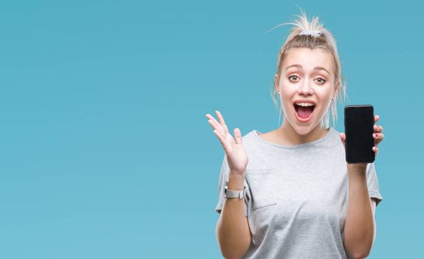 Young blonde woman showing screen of smartphone over isolated background very happy and excited, winner expression celebrating victory screaming with big smile and raised hands - Photo, Image