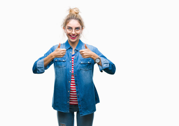 Young beautiful blonde woman wearing glasses over isolated background success sign doing positive gesture with hand, thumbs up smiling and happy. Looking at the camera with cheerful expression, winner gesture. - 写真・画像
