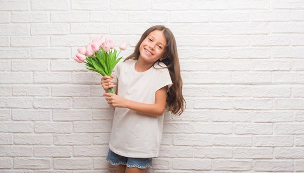 Молода іспаномовна дитина над білою цегляною стіною тримає квіти в день матері з щасливим обличчям, що стоїть і посміхається з впевненою посмішкою, показуючи зуби
 - Фото, зображення