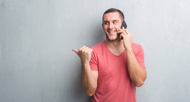 Young caucasian man over grey grunge wall talking on the phone pointing and showing with thumb up to the side with happy face smiling - Φωτογραφία, εικόνα