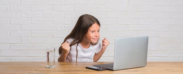 Nuori latino lapsi istuu pöydällä tietokoneen kannettava tietokone huutaa ylpeä ja juhlii voittoa ja menestystä erittäin innoissaan, hurraavat tunteita
 - Valokuva, kuva