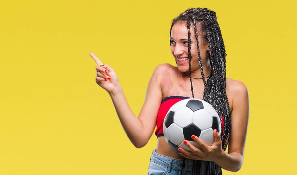 Giovane capelli intrecciati afroamericano con marchio di nascita tenendo palla da calcio su sfondo isolato molto felice puntando con mano e dito di lato
 - Foto, immagini