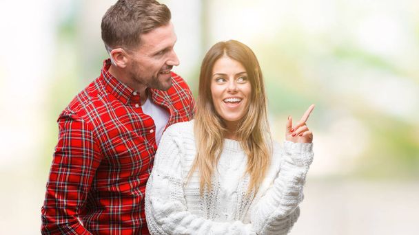 Jeune couple amoureux portant un pull d'hiver sur fond isolé avec un grand sourire sur le visage, pointant avec la main et le doigt vers le côté regardant la caméra
. - Photo, image