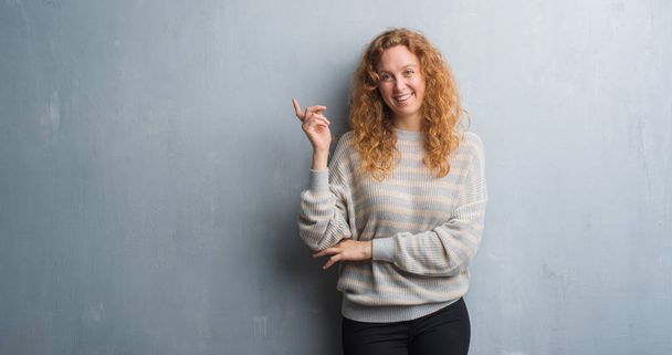Young redhead woman over grey grunge wall with a big smile on face, pointing with hand and finger to the side looking at the camera. - Foto, Imagen