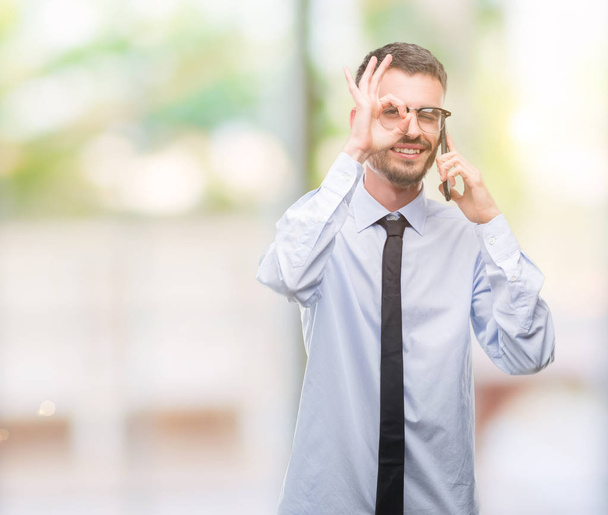 Jeune homme d'affaires adulte parlant au téléphone avec le visage heureux souriant faire ok signe avec la main sur les yeux en regardant à travers les doigts
 - Photo, image
