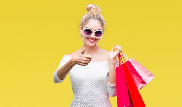 Young beautiful blonde woman holding shopping bags over isolated background happy with big smile doing ok sign, thumb up with fingers, excellent sign - Fotografie, Obrázek