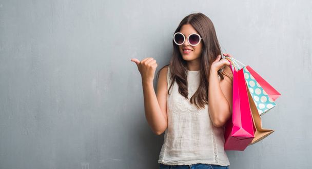 Fiatal barna nő grunge szürke falon gazdaság színes bevásárló táskák, mutató és hüvelykujjával, akár a boldog arc mosolyogva oldalon mutatja - Fotó, kép