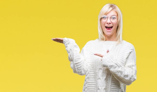 Young beautiful blonde woman wearing winter sweater and glasses over isolated background amazed and smiling to the camera while presenting with hand and pointing with finger. - Photo, Image