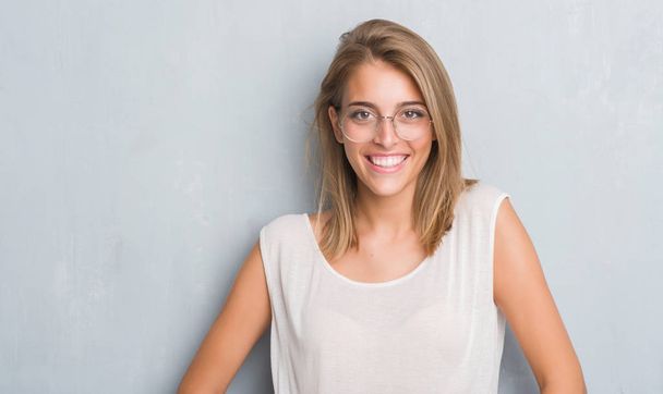 Όμορφη νεαρή γυναίκα που στέκεται πάνω από το τοίχο γκρι grunge φορώντας γυαλιά με ένα χαρούμενο πρόσωπο στέκονται και να χαμογελούν με αυτοπεποίθηση χαμόγελο δείχνει τα δόντια - Φωτογραφία, εικόνα