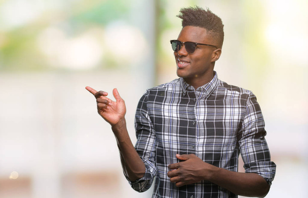 junger afrikanisch-amerikanischer Mann trägt Sonnenbrille über isoliertem Hintergrund mit einem breiten Lächeln im Gesicht und zeigt mit der Hand und dem Finger zur Seite in die Kamera. - Foto, Bild