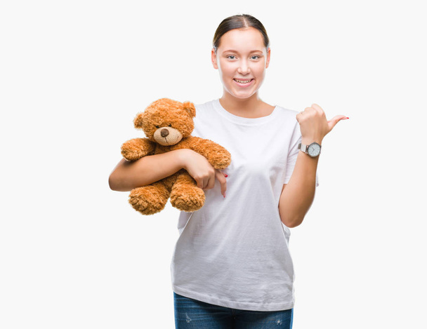 Young caucasian woman holding teddy bear over isolated background pointing and showing with thumb up to the side with happy face smiling - 写真・画像